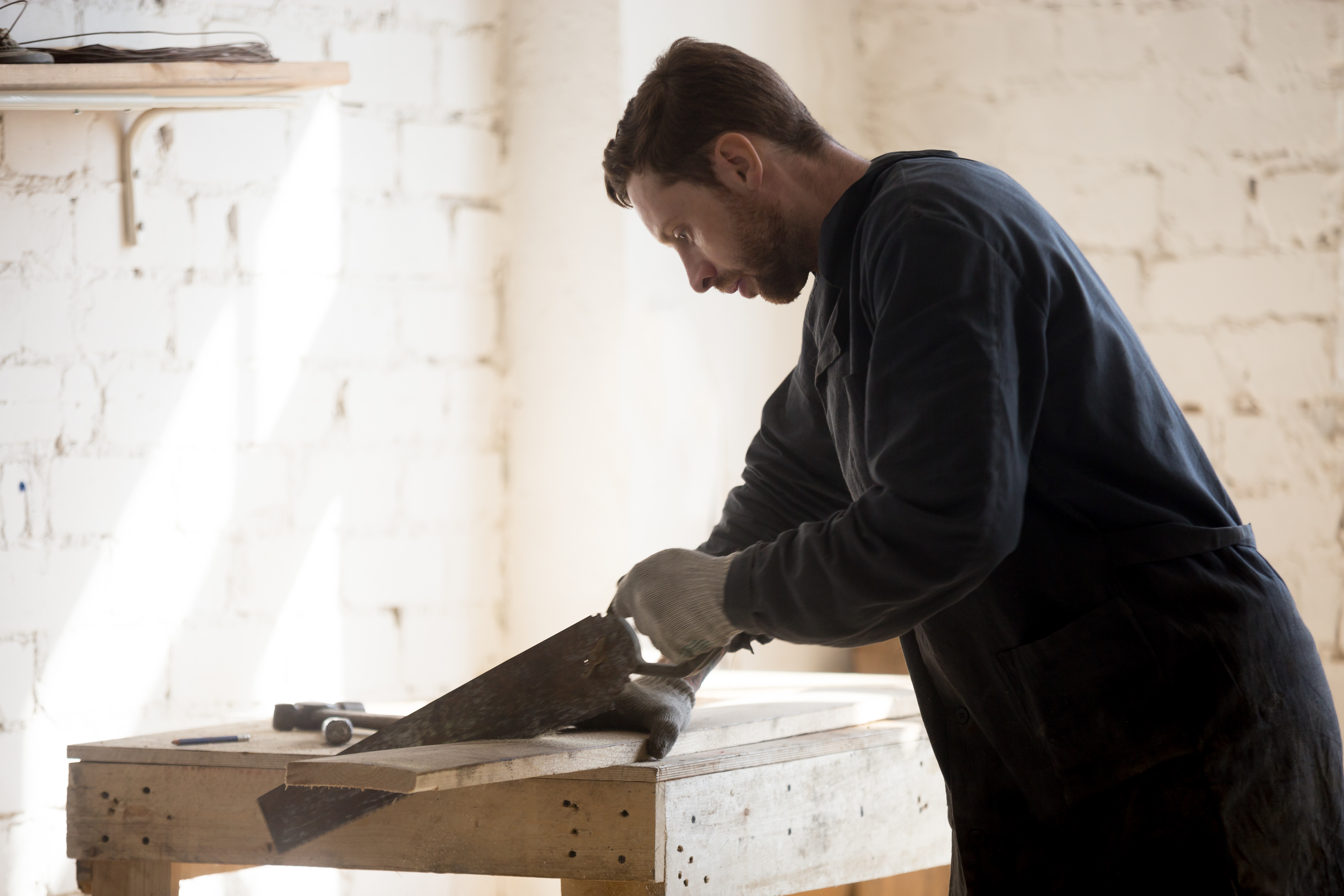 Enthusiast passionate about woodworking hobby cutting wooden board with old-fashioned hand saw on workbench at workshop. Restorer of antique furniture working at new project. Home based business idea